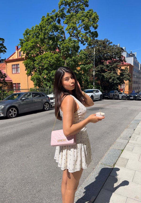 Ung kvinde i hvid kjole med polkaprikker, der holder en iskremkop, står på en solrig gade med parkerede biler og træer bag sig.