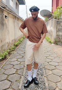 T-shirt marron à manches courtes en coton avec une coupe décontractée, associée à un short crème arrivant aux genoux, des chaussettes blanches et des chaussures noires sans lacets.