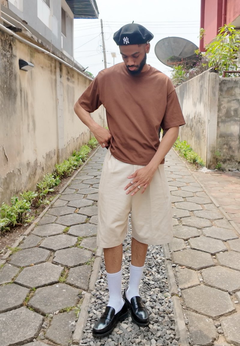 T-shirt marron à manches courtes en coton avec une coupe décontractée, associée à un short crème arrivant aux genoux, des chaussettes blanches et des chaussures noires sans lacets.