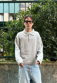 Pull gris tricoté avec col, patte de boutonnage à trois boutons et ourlet côtelé, associé à un pantalon en denim bleu clair. Feuillage vert en arrière-plan.