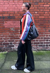 Chaqueta de gran tamaño en colores vibrantes con rayas moradas, rojas y azules, combinada con pantalones anchos negros y zapatillas blancas. Se presenta un bolso negro.