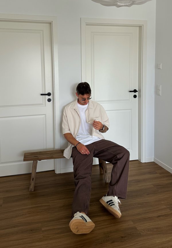 Uomo con occhiali da sole seduto su una panchina di legno in una stanza con pareti bianche e due porte bianche chiuse.