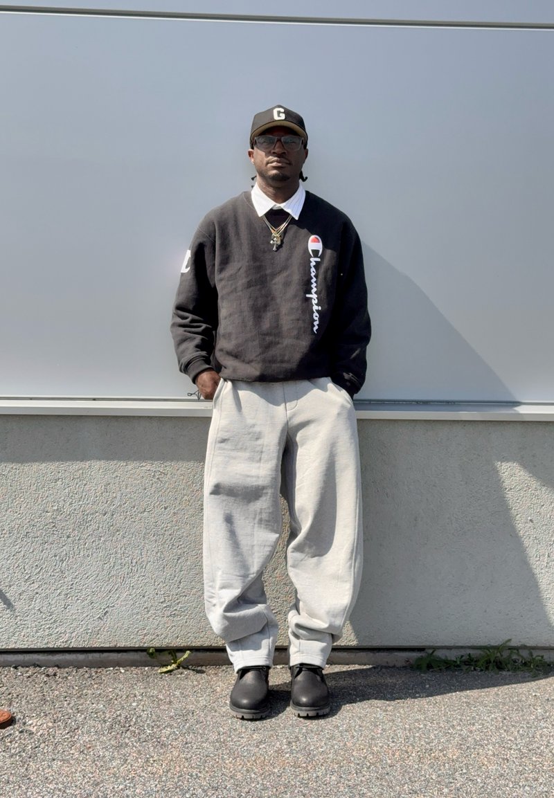 Personne portant un sweat Champion noir, un col de chemise blanc, un pantalon ample gris clair, des bottes noires, une casquette noire et des lunettes de soleil, debout contre un mur gris.