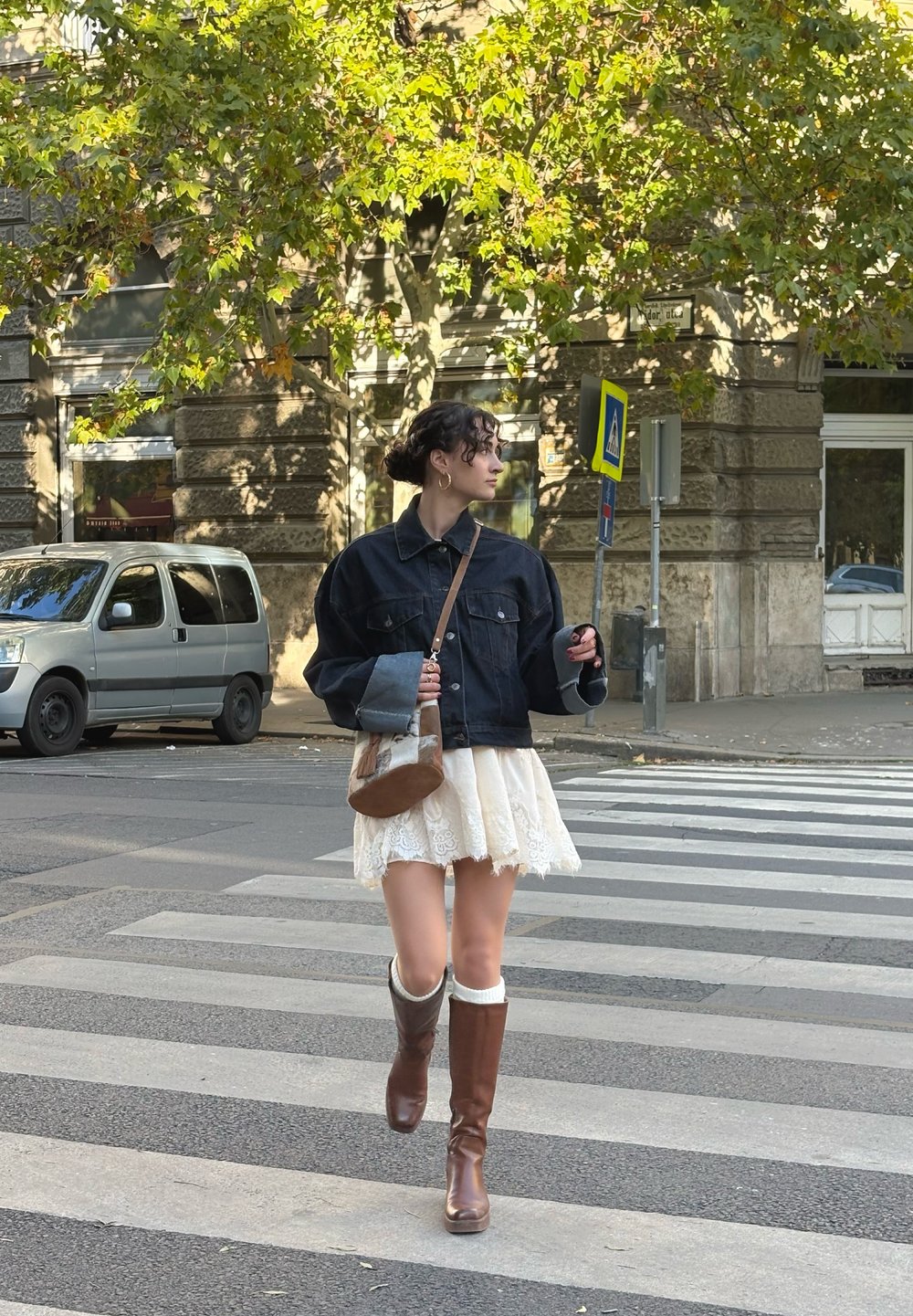 Casaco de ganga com corte oversized, saia de renda creme, botas castanhas até ao joelho e uma pequena mala de ombro castanha, a caminhar num passamento.