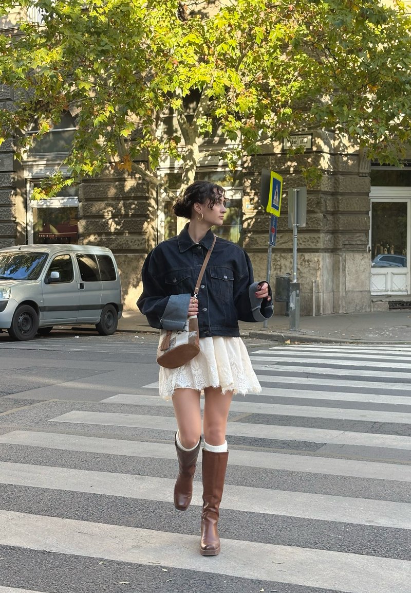 Denim jacket with oversized fit, cream lace skirt, brown knee-high boots, and a small brown shoulder bag, walking on a crosswalk.