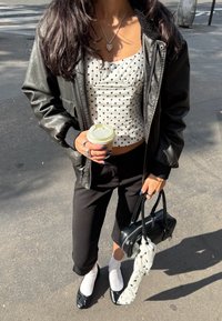 Mustat nahkabomberitakki, valkoinen pilkullinen crop-top, mustat kapenevat housut, mustat litteät kengät, valkoiset sukat, pieni musta laukku, käsissä kahvikuppi.