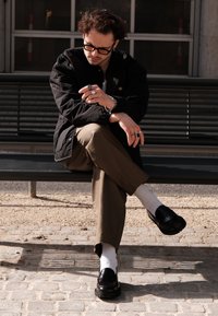 Casaco acolchoado preto, camisa branca, calças olivais e mocassins de couro preto com solas texturizadas. Vários anéis e pulseiras de prata em destaque.
