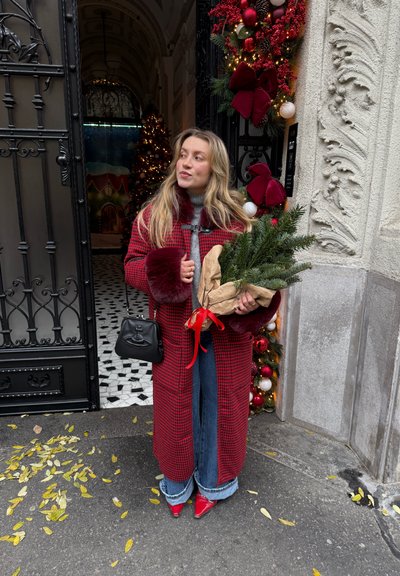 Abrigo rojo a cuadros con puños de piel sintética, suéter gris, vaqueros azules de pierna ancha, zapatos rojos puntiagudos, sosteniendo ramas verdes envueltas con una cinta roja. Bolso negro.