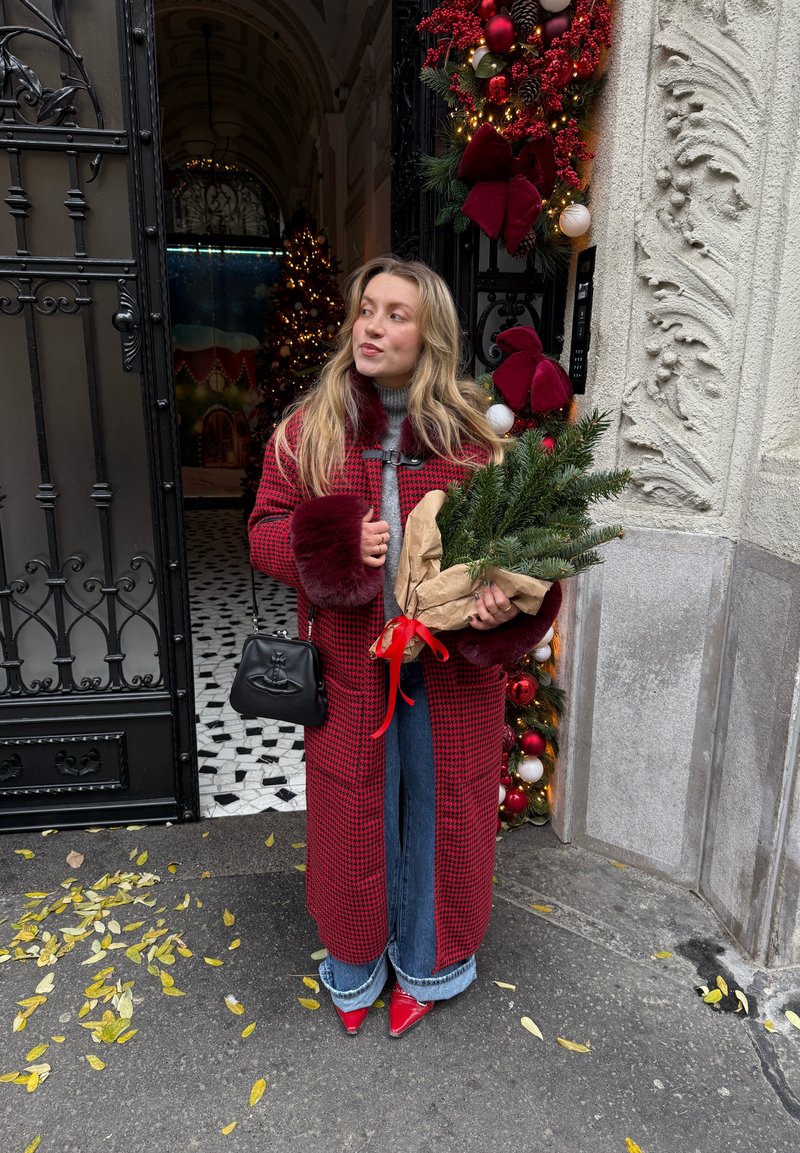 Manteau à carreaux rouge avec poignets en fausse fourrure, pull gris, jean large bleu, chaussures rouges pointues, tenant un bouquet de verdure enveloppé avec un ruban rouge. Sac à main noir.