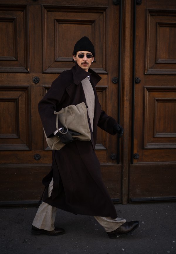 Homme en long manteau foncé, pantalon beige et bonnet noir marchant devant une porte en bois, portant un grand sac beige, des lunettes de soleil et des gants.