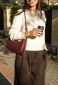 Bílý svetr na zip, hnědé široké kalhoty, burgundy kožená kabelka přes rameno, držící černý hrnek s kávou, venkovní prostředí se zelení.
