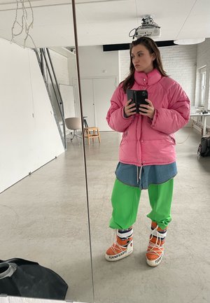 Chaqueta rosa esponjosa, camisa de mezclilla, pantalones verdes brillantes y zapatillas naranjas con acentos blancos. Reflexión en el espejo, fondo de estudio minimalista.