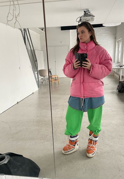 Chaqueta rosa esponjosa, camisa de mezclilla, pantalones verdes brillantes y zapatillas naranjas con acentos blancos. Reflexión en el espejo, fondo de estudio minimalista.