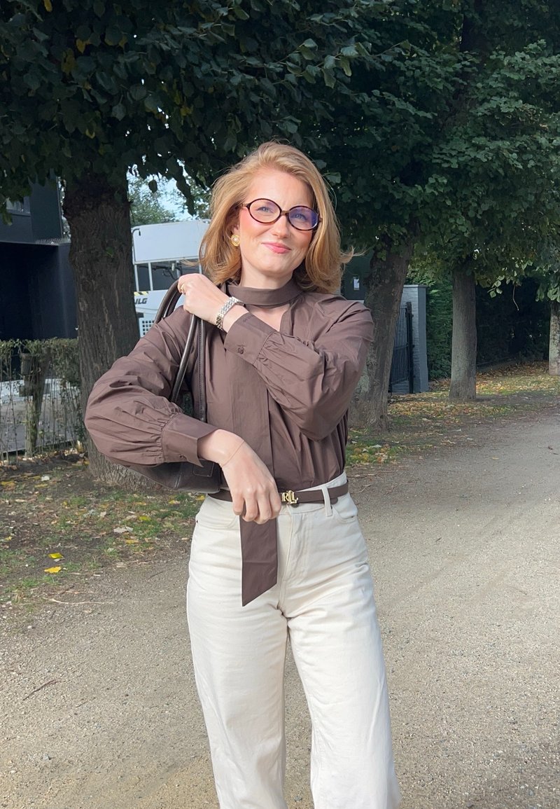 Bluse mit langen Ärmeln in Braun und einem Halstuch, kombiniert mit kremfarbigen, weiten Hosen. Accessoires in Form einer dunklen Handtasche und übergroßen Brillen.