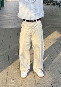 Pantaloni a gamba larga di un beige chiaro, realizzati in un tessuto dalla texture liscia, abbinati a sneaker bianche. In piedi su un pavimento testurizzato.