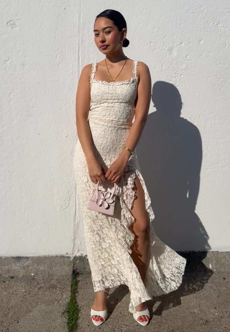 Jeune femme en robe blanche en dentelle avec une fente sur la cuisse, tenant un petit sac à main rose, debout contre un mur blanc ensoleillé.