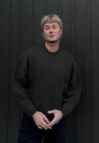 Pull en tricot vert foncé avec un col rond, des épaules tombantes et des poignets côtelés, associé à un pantalon foncé sur un fond texturé.