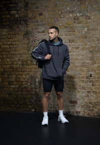 Γκρίζα φούτερ με καγκουρό τσέπη, λευκές πλευρικές ρίγες, συνδυασμένο με μαύρες βερμούδες. Λευκά αθλητικά παπούτσια και μια μαύρη τσάντα πλάτης ολοκληρώνουν την εμφάνιση.