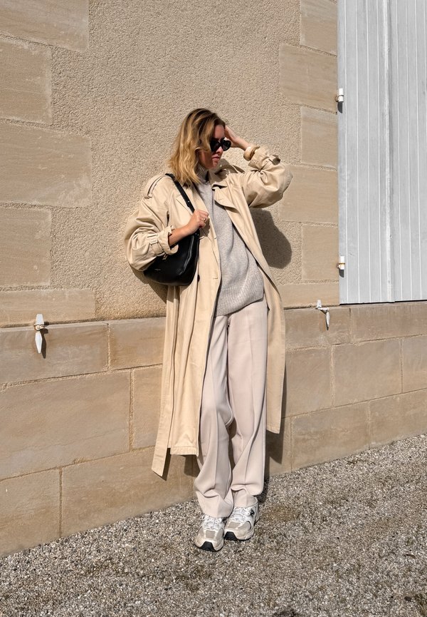 Beige trenchcoat, grå genser, beige vide bukser og sølvfargede sneakers. Svart håndveske og solbriller. Teksturert vegg bakgrunn.
