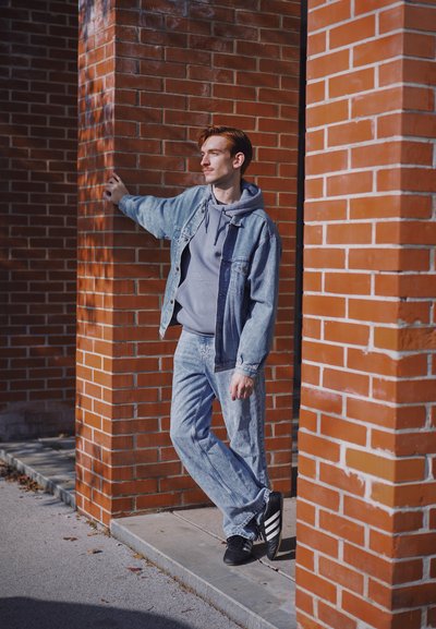 Chaqueta vaquera en tonos azules sobre una sudadera gris, combinada con vaqueros azul claro y zapatillas negras con rayas blancas, frente a una pared de ladrillo.