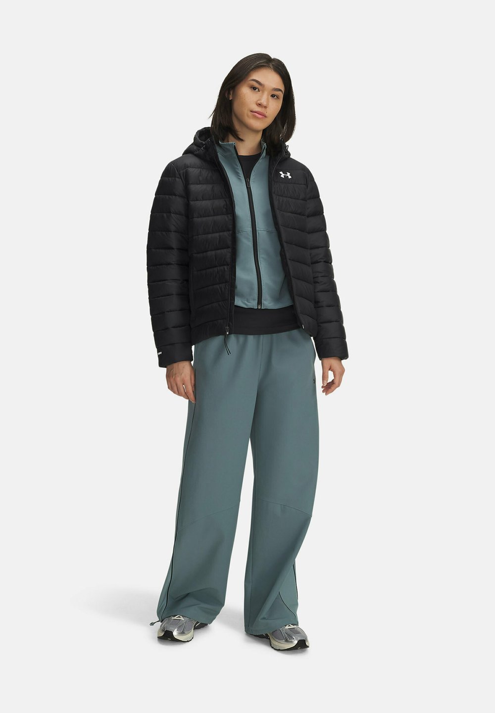 Zwarte pufferjack met horizontale stiksels, teal rits-hoodie eronder, en brede teal broek. Grijze sneakers maken de outfit af.