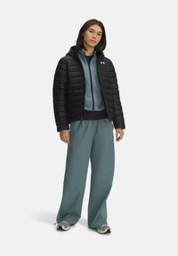 Zwarte pufferjack met horizontale stiksels, teal rits-hoodie eronder, en brede teal broek. Grijze sneakers maken de outfit af.