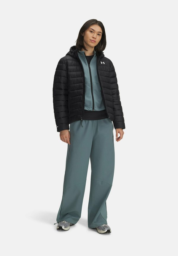 Zwarte pufferjack met horizontale stiksels, teal rits-hoodie eronder, en brede teal broek. Grijze sneakers maken de outfit af.