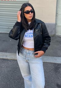 Giubbotto bomber in pelle nera con tasca a zip, T-shirt bianca corta con stampa blu e jeans a gamba larga in denim chiaro. La modella indossa occhiali da sole.