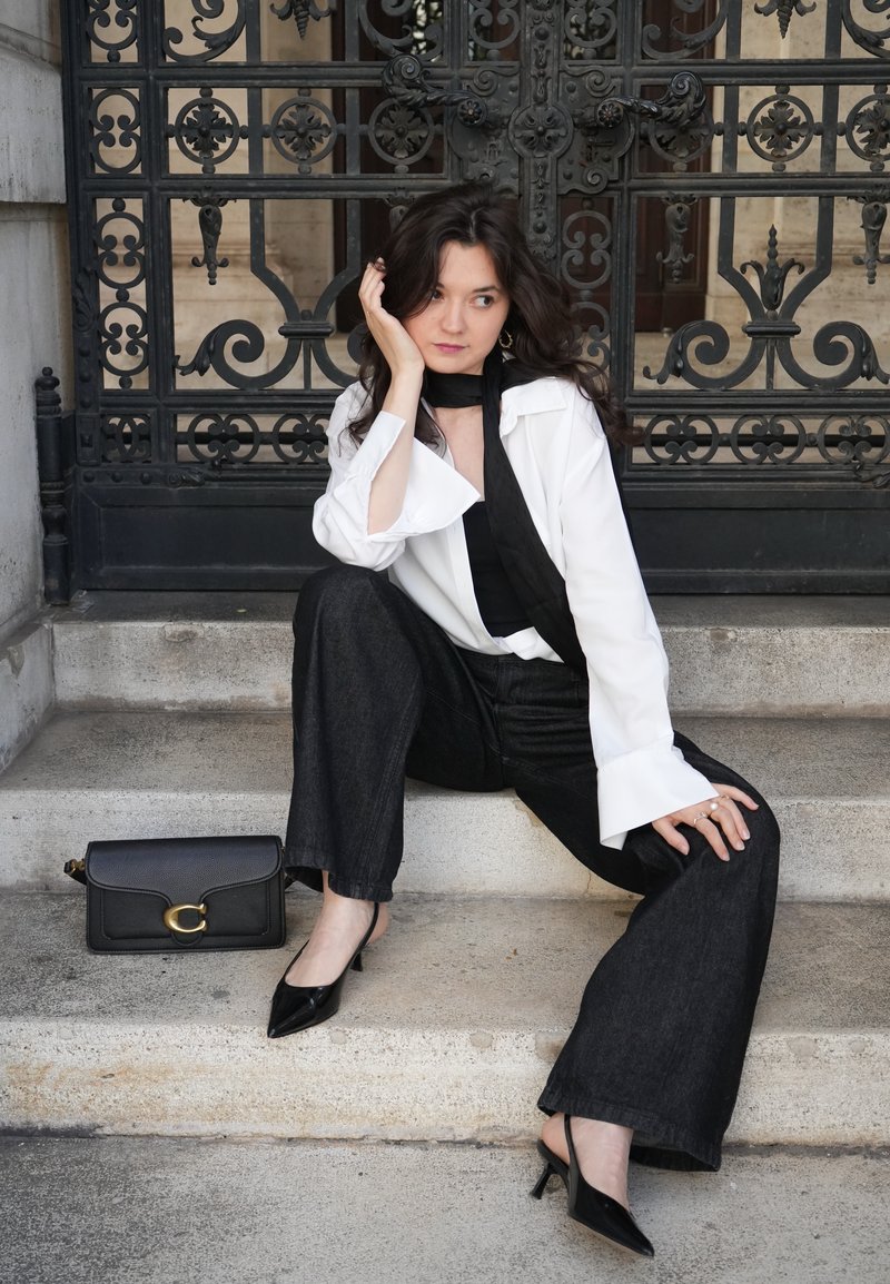 Femme aux cheveux foncés portant une chemise blanche, un pantalon noir et des talons, assise sur des marches en pierre à côté d'un sac à main noir, devant une porte en fer ornée.
