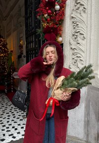 Manteau pied-de-poule rouge et noir avec une capuche en fausse fourrure bordeaux, tenant une petite branche de pin enveloppée dans du papier brun avec un ruban rouge.