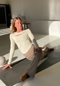 Top de manga comprida creme com detalhe recortado, combinado com calças flare castanho claro. Em um tapete de yoga escuro em um espaço minimalista iluminado pelo sol.