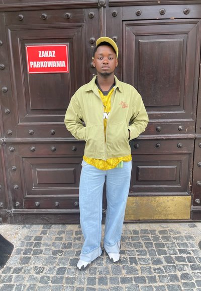 Chaqueta de pana verde sobre una camisa amarilla estampada, combinada con unos jeans de pierna ancha de color azul claro y unos zapatos de color claro. El fondo presenta una puerta de madera.