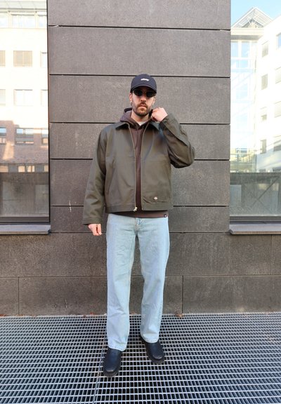 Hombre con gorra negra, gafas de sol, chaqueta verde oscuro sobre sudadera marrón, vaqueros azul claro y zapatos negros, de pie contra una pared gris al aire libre.