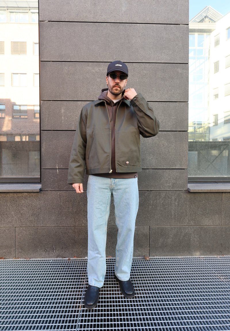 Homme portant une casquette noire, des lunettes de soleil, une veste vert foncé par-dessus un sweat à capuche marron, un jean bleu clair et des chaussures noires, debout contre un mur gris en plein air.