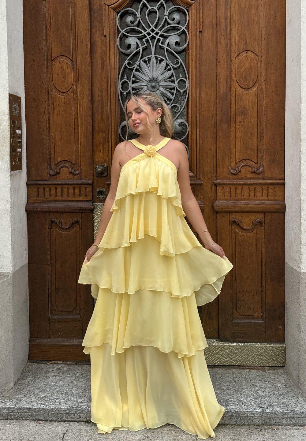Gelbes, mehrlagiges Chiffonkleid mit Halterneck-Design, das Rüschen und eine florale Akzentuierung am Ausschnitt aufweist, vor einem Holztür-Hintergrund.