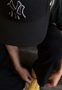 Zwarte pet met een wit "NY"-logo, zichtbare stiksels en een vlakke klep. Gele schoenen deels zichtbaar op een gestructureerd oppervlak.