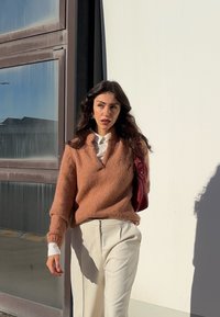 Sweter w kolorze różowym z dekoltem w serek, o miękkiej fakturze, w połączeniu z białą koszulą i kremowymi spodniami; trzyma burgundową torbę.