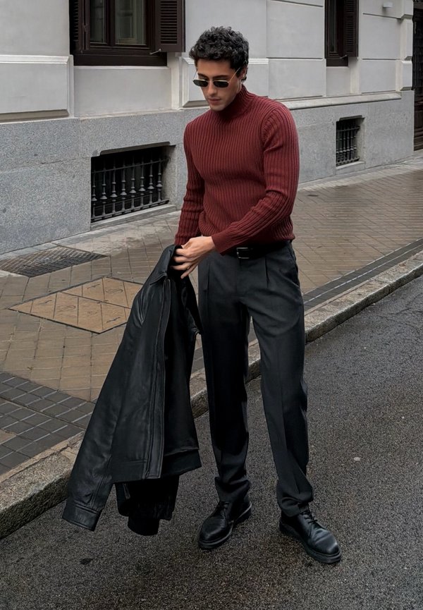 Hombre vistiendo un suéter de canalé color burdeos y pantalones negros sostiene una chaqueta de cuero negra mientras está de pie en la acera de una ciudad cerca de un edificio.