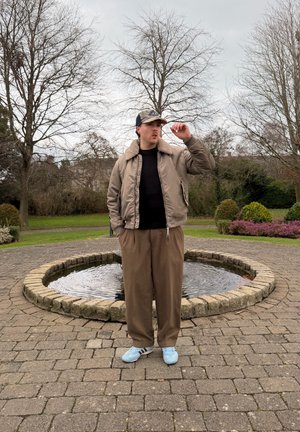 Joven con chaqueta marrón, pantalones y zapatillas deportivas azules ajustando una gorra, de pie en un círculo pavimentado alrededor de una pequeña fuente de agua en un parque.
