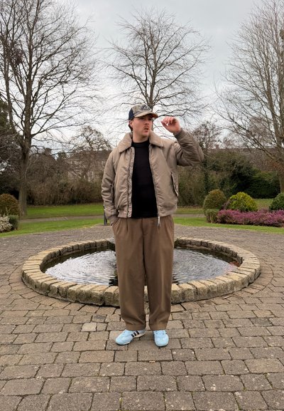 Joven con chaqueta marrón, pantalones y zapatillas deportivas azules ajustando una gorra, de pie en un círculo pavimentado alrededor de una pequeña fuente de agua en un parque.