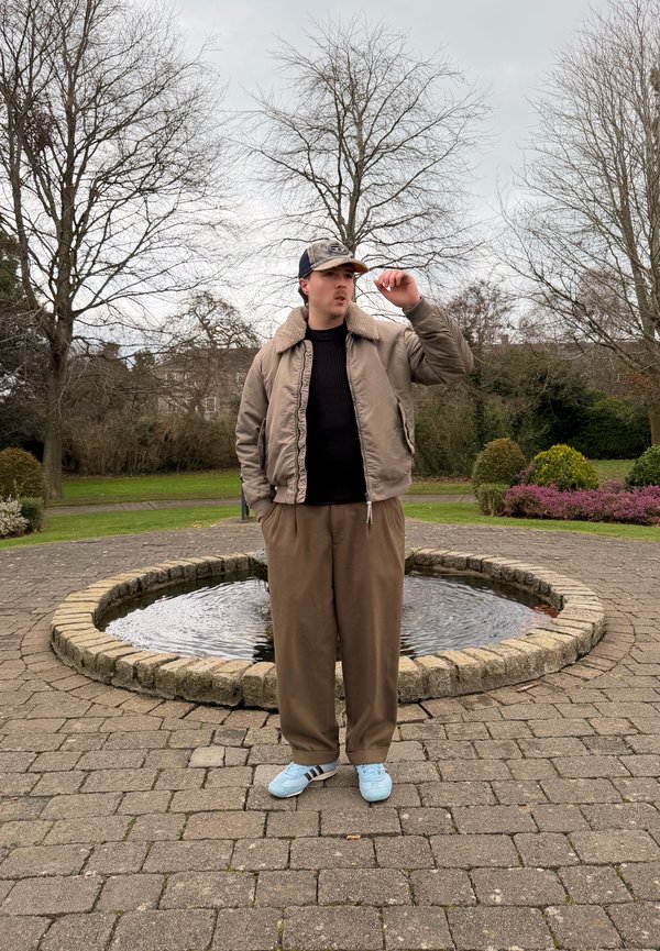 Νεαρός άνδρας με καφέ μπουφάν, παντελόνια και μπλε αθλητικά παπούτσια προσαρμόζει το καπέλο του, στέκεται σε πλακόστρωτο κύκλο γύρω από ένα μικρό συντριβάνι σε πάρκο.