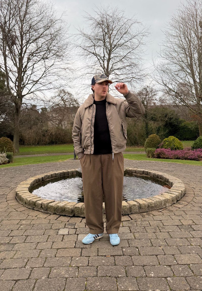 Joven con chaqueta marrón, pantalones y zapatillas deportivas azules ajustando una gorra, de pie en un círculo pavimentado alrededor de una pequeña fuente de agua en un parque.
