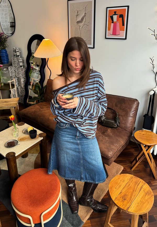 Woman in a striped off-shoulder top and denim skirt holding a cup, standing in a cosy living room with a brown sofa and wooden stools.