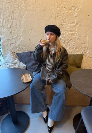 Joven mujer con boina negra y chaqueta de cuero bebe café sentada entre dos mesas en un café con pared beige texturizada de fondo.