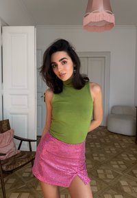 Haut vert sans manches associé à une mini-jupe rose à sequins avec une fente latérale. Pièce avec un sol en bois clair et un luminaire.