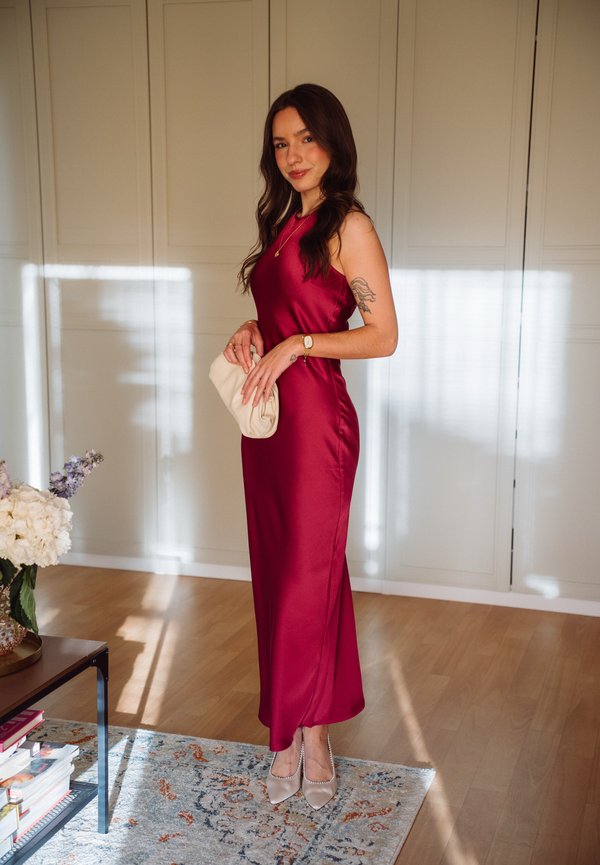 Femme portant une robe rouge en satin sans manches et des talons beiges, tenant une pochette beige, debout à l'intérieur près d'un tapis et d'une table avec des fleurs.