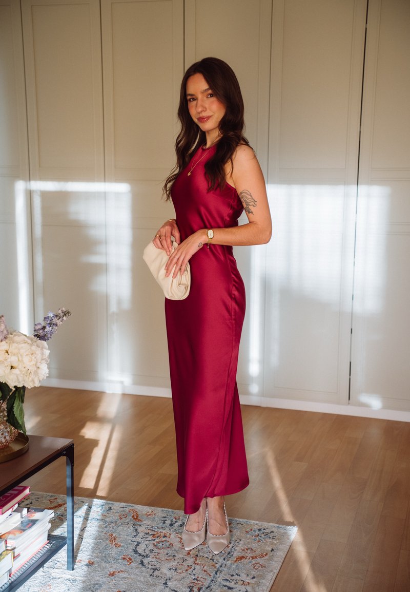 Femme portant une robe rouge en satin sans manches et des talons beiges, tenant une pochette beige, debout à l'intérieur près d'un tapis et d'une table avec des fleurs.