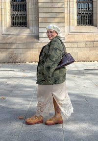 Jachetă cu model camuflaj peste o fustă de satin crem cu dantelă la margine. Sandale cu platformă maro. Geantă maro. Cadru urban în aer liber.
