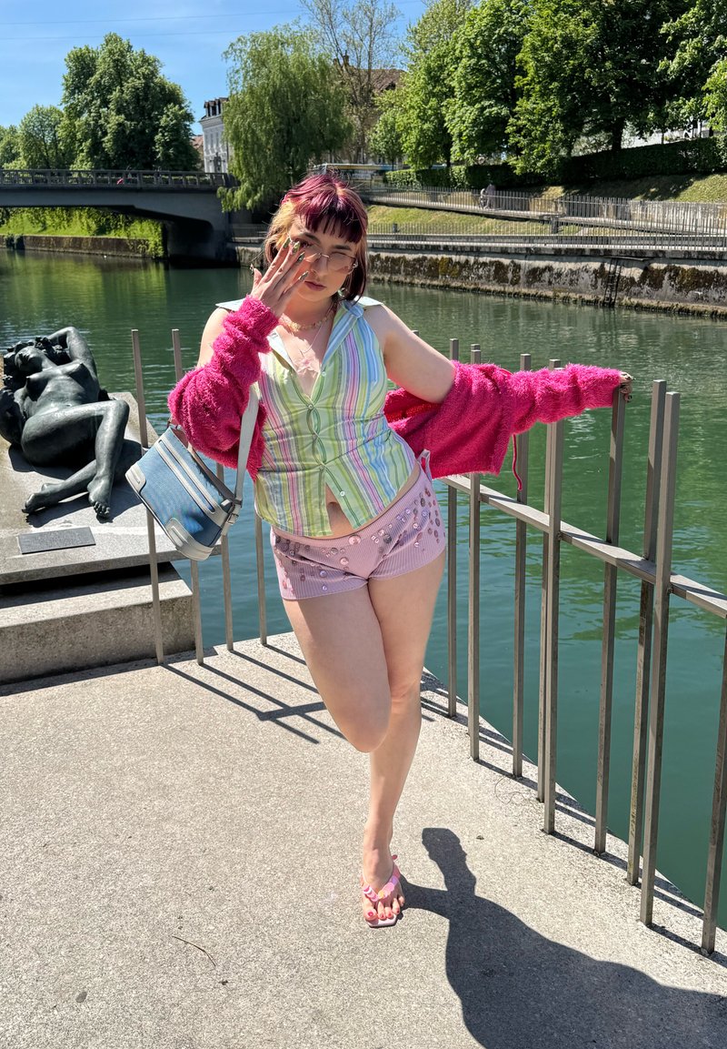 Jeune femme avec des cheveux teints portant un débardeur rayé, un short rose et un cardigan, posant près d'une rambarde à côté d'une sculpture de rivière par une journée ensoleillée.