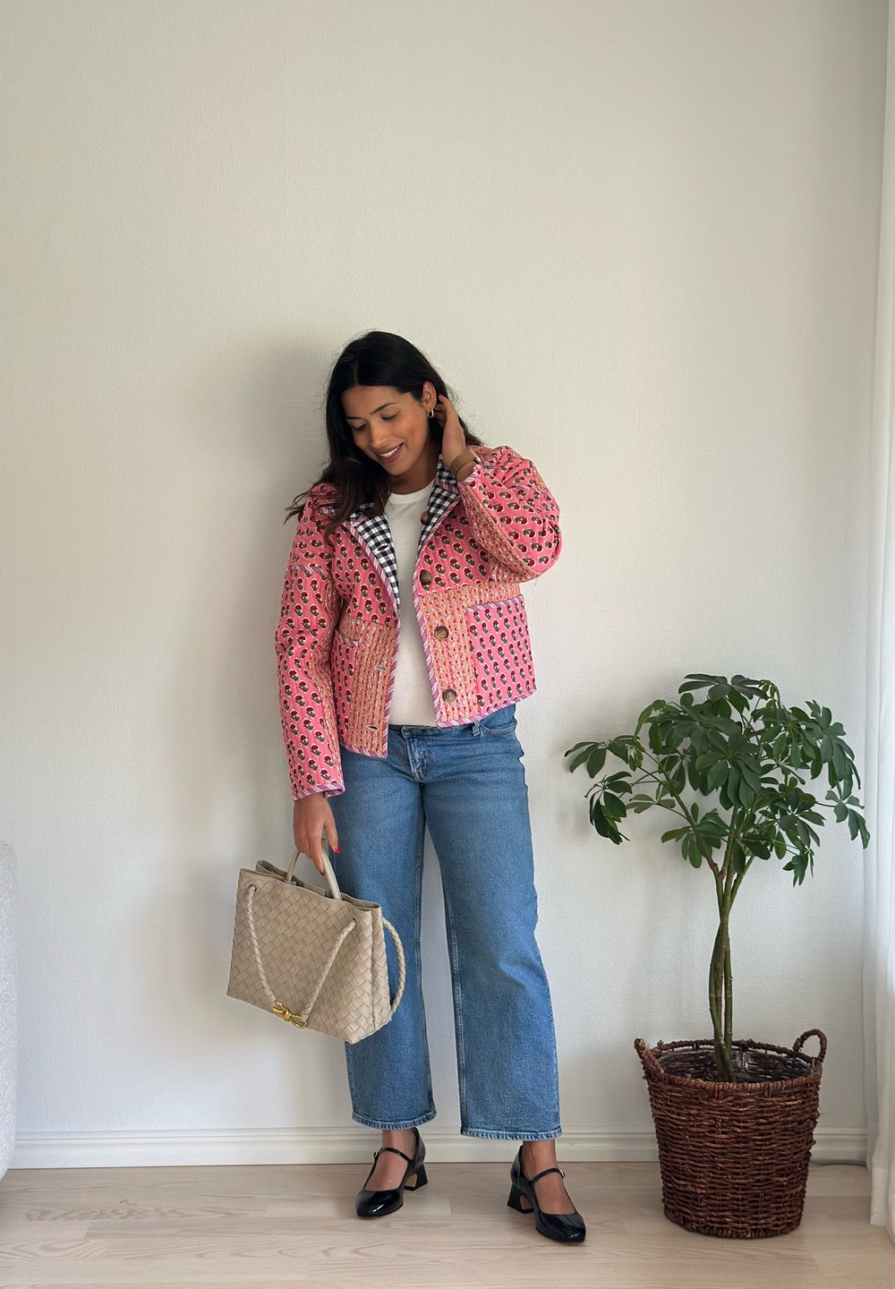Rosa gemusterte Jacke mit schwarzem kariertem Kragen, kombiniert mit blauen Jeans und schwarzen Schuhen. Hält eine beige gewebte Handtasche. Innenraum.